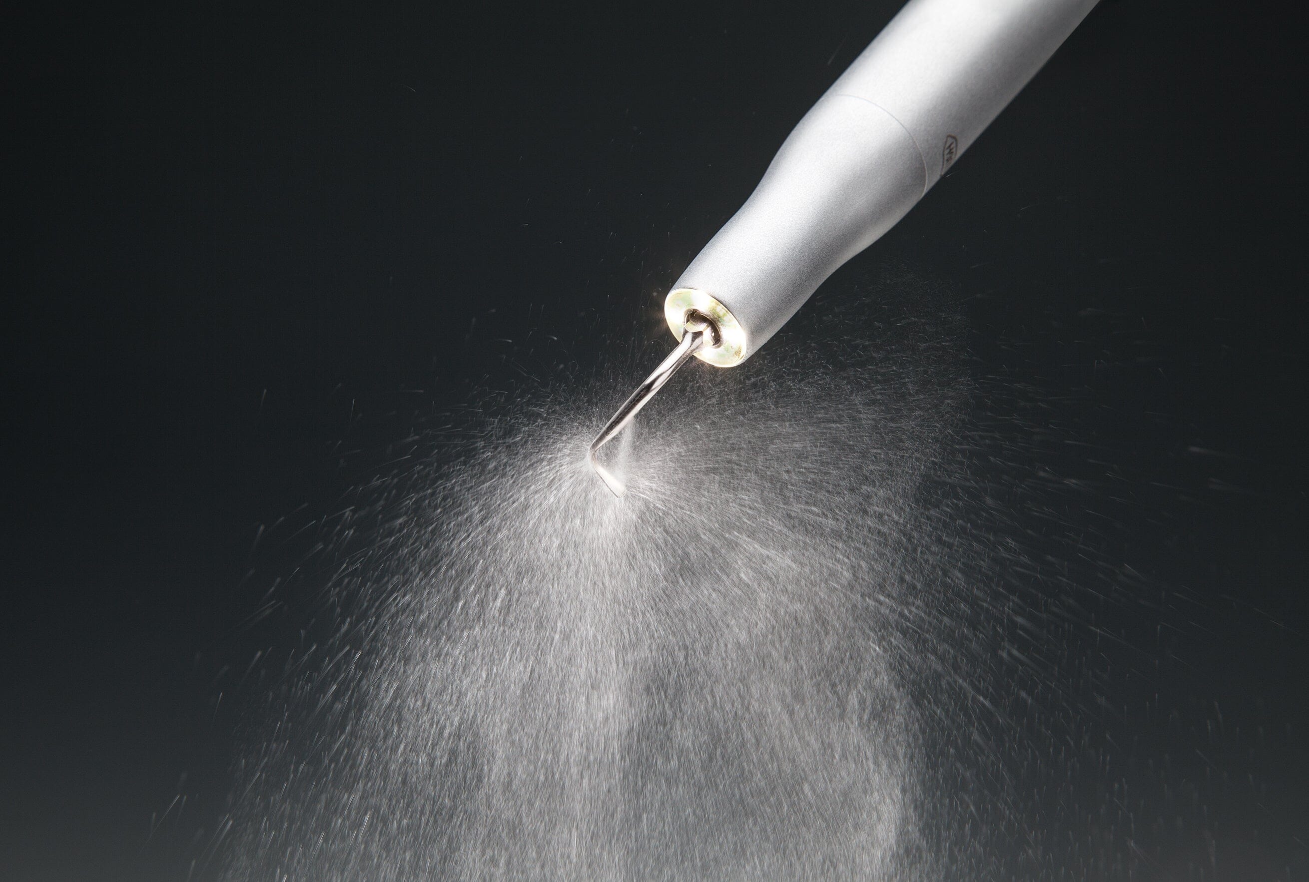 A close-up of the W&H Piezomed SA-320 Handpiece with Cable emitting a fine mist of water spray during ultrasonic bone surgery, set against a dark background.