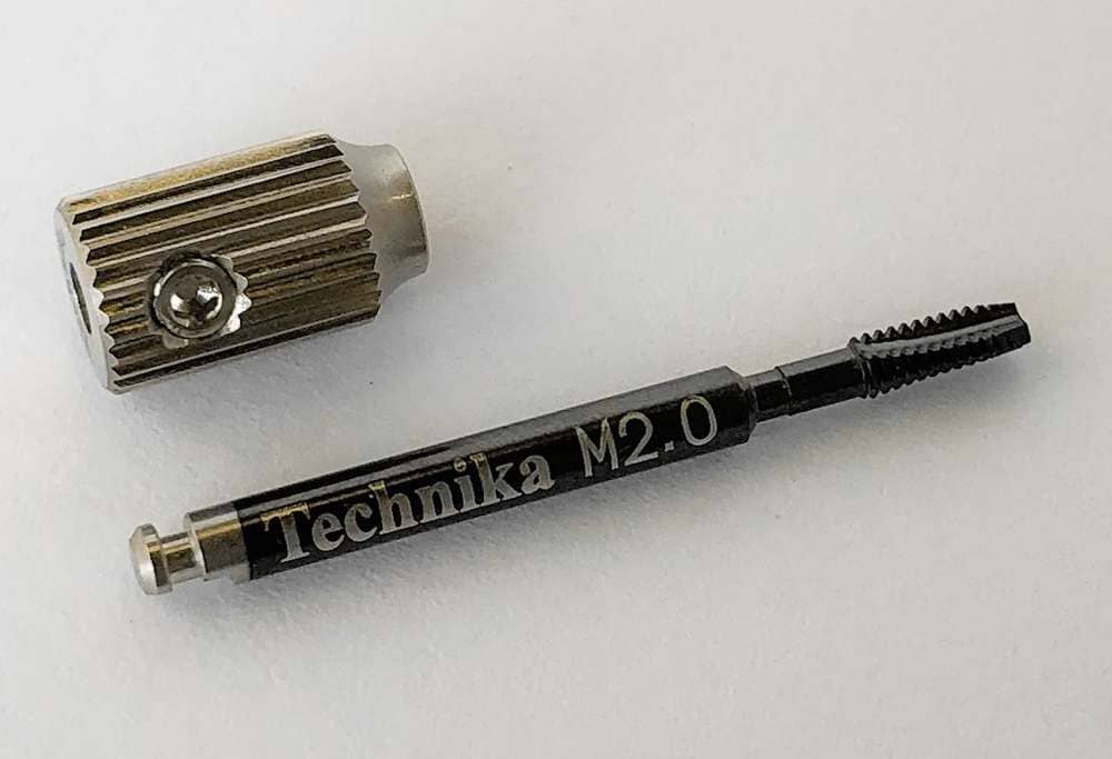 A Dentech Technika Repair Tap with Handle — M1.6 & M2.0, used for internal implant thread repair, is shown on a white surface, featuring a textured metal cap and threaded tip.