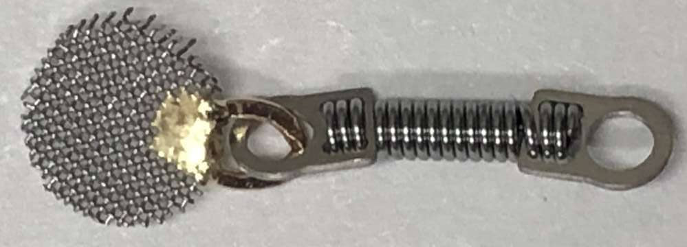The Xemax Cusp-Lok Bracket Titanium Spring with Mesh is a small metal device featuring a circular mesh top, a central titanium spring, and two flat end loops. The mesh is secured with a spot of yellowish material.