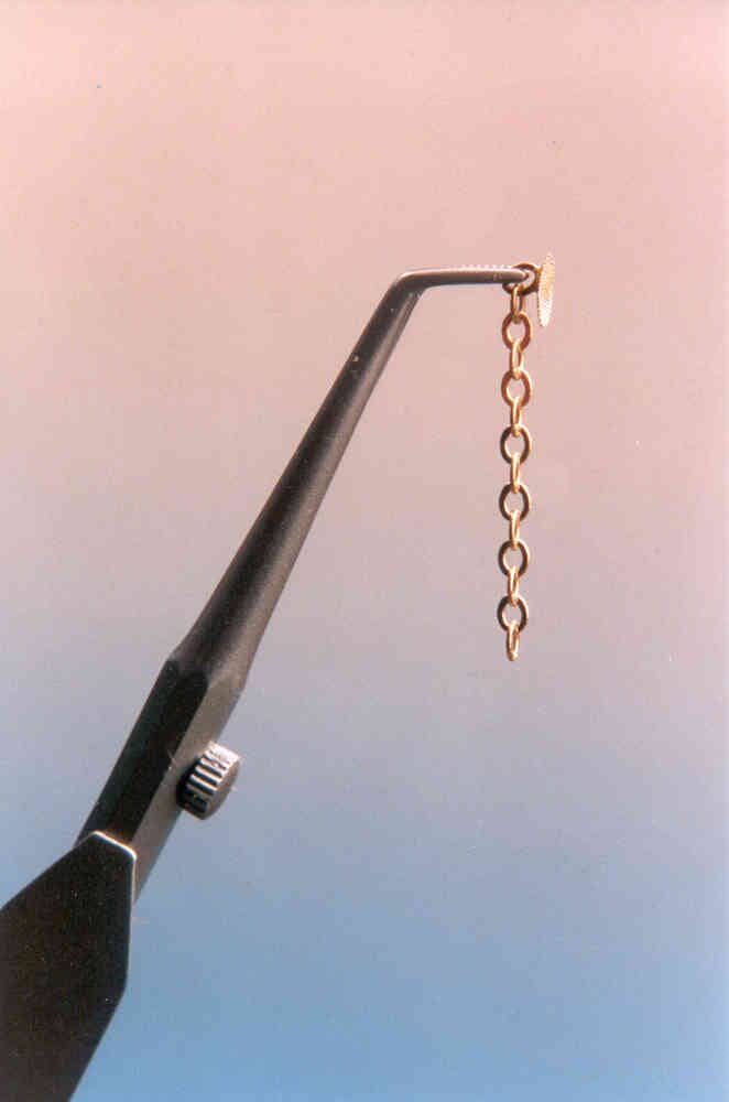 A black tweezer holds a delicate Xemax Cusp-Lok Chain & Mesh 14K Gold orthodontic traction system, featuring oval links and suspended vertically against a pink-to-blue gradient background.