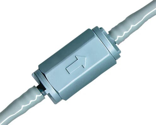 A close-up of a Quality Aspirators Backflow Valve for Saliva Ejector Tubing, showing its metallic cylindrical body with a hexagonal tightening section, an arrow for flow direction, and tubing connected at both ends.